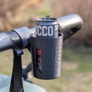 Könnte beinhalten: Ein schwarzer Metallbecherhalter, der an einem schwarzen Lenker befestigt ist und eine Dose "ROCCO NO"-Getränk hält. Der Becherhalter hat eine strukturierte Oberfläche und ein Logo. Ein schwarzer Riemen mit weißem Text hängt vom Lenker.