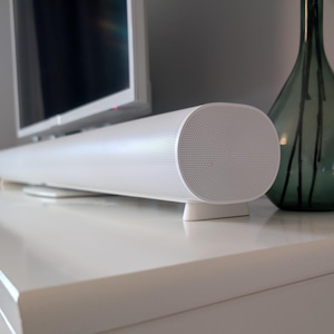 以下が含まれることがあります： 前面にスピーカーグリルが付いた白いサウンドバー。サウンドバーは、フラットスクリーンテレビの前にある白い表面の上に置かれています。