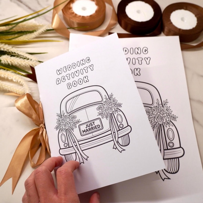 May include: Two white "Wedding Activity Book" booklets with a black and white illustration of a car with "Just Married" on the license plate. The booklets are decorated with floral arrangements and ribbons.