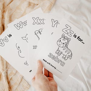 Pu&ograve; includere: Una mano tiene in mano un mucchio di schede dell'alfabeto bianche. Ogni scheda presenta una lettera e un'illustrazione corrispondente, come una zebra per la lettera Z. Il testo "made with love by" &egrave; stampato in basso su ogni scheda.