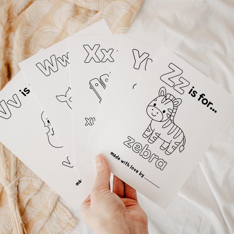 Pu&ograve; includere: Una mano tiene in mano un mucchio di schede dell'alfabeto bianche. Ogni scheda presenta una lettera e un'illustrazione corrispondente, come una zebra per la lettera Z. Il testo "made with love by" &egrave; stampato in basso su ogni scheda.