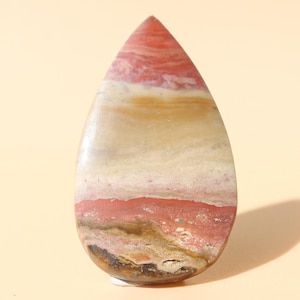 Puede incluir: Una gema pulida en forma de lágrima. La piedra presenta capas de colores rojo, blanco, beige y marrón. La gema se encuentra sobre un fondo beige claro.