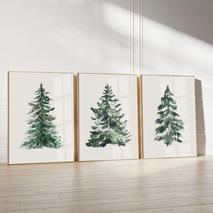 May include: Three framed art prints featuring watercolor paintings of evergreen trees. The trees are rendered in shades of green and are set against a cream-colored background. The frames have a gold finish, and the artwork is displayed on a wooden floor.