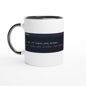 Op de afbeelding: Een witte keramische mok met een zwarte binnenkant en handvat. De voorzijde toont een donkere terminalvenster-afbeelding met de tekst: $ rm -rf hopes_and_dreams, gevolgd door de foutmelding: rm: hopes_and_dreams: not found.