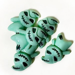 May include: A collection of light teal skull-shaped beads with black stitched details around the eyes and mouth. The beads have a glossy finish and are arranged on a white background. These Halloween-themed beads are ideal for crafting.