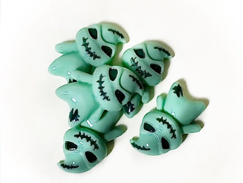 May include: A collection of light teal skull-shaped beads with black stitched details around the eyes and mouth. The beads have a glossy finish and are arranged on a white background. These Halloween-themed beads are ideal for crafting.
