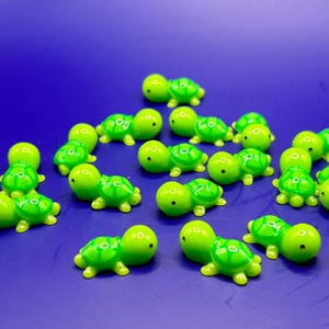 Può includere: Un gruppo di 15 piccole figurine di tartarughe di plastica verde con occhi neri. Le tartarughe sono disposte in modo casuale su una superficie blu.