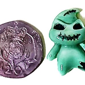 May include: A green figurine of a character with black eyes and a stitched mouth, next to a Twenty Pence coin. The figurine has a stitched appearance and a rounded shape. The coin is silver and shows a crown.