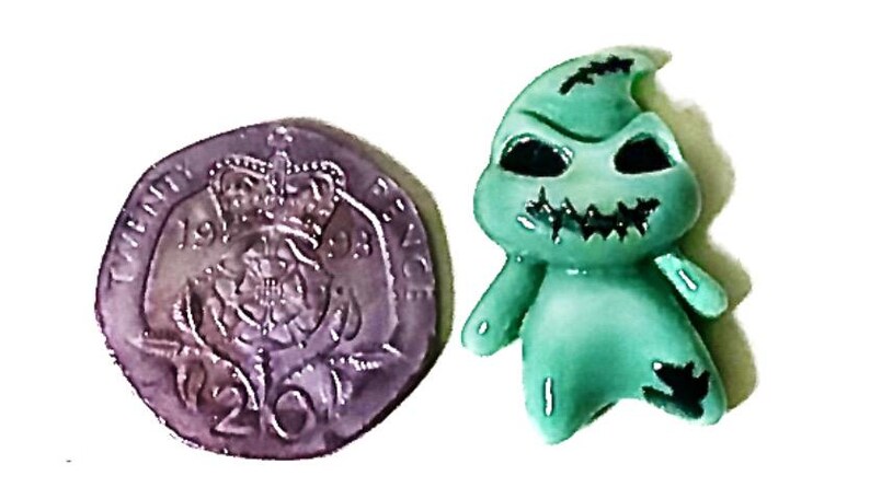 May include: A green figurine of a character with black eyes and a stitched mouth, next to a Twenty Pence coin. The figurine has a stitched appearance and a rounded shape. The coin is silver and shows a crown.
