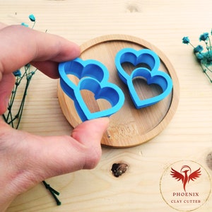 Può includere: Due tagliabiscotti a forma di cuore in plastica blu, uno impilato sull'altro, su una superficie di legno.