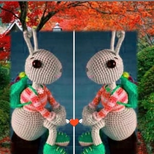 Puede incluir: Dos peluches de conejo de crochet gris con camisas a rayas rojas y blancas y mochilas verdes. Los conejos están sentados uno frente al otro con las piernas cruzadas. Están frente a un fondo borroso de follaje otoñal.