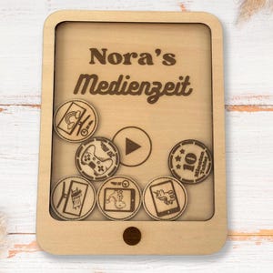 Könnte beinhalten: Eine Holzkiste in Form eines Telefons mit einem transparenten Plastikfenster. Die Kiste hat den Text "Nora's Medienzeit" auf der Vorderseite eingraviert. Das Fenster zeigt verschiedene kleine Holztoken mit unterschiedlichen Symbolen, darunter ein Fernseher, ein Gamecontroller und ein Einhorn.