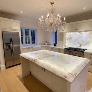 May include: A luxurious kitchen with a large island featuring a white and gold marble countertop. The kitchen includes stainless steel appliances, white cabinetry, and a crystal chandelier. The flooring is light-colored wood.