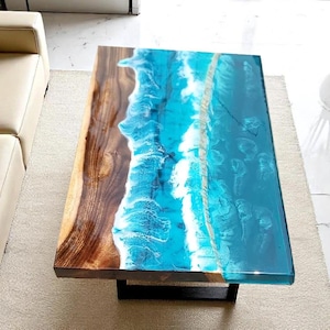 May include: A rectangular coffee table with a unique design. The table features a wooden surface with a brown finish on one side and a blue and white resin design resembling ocean waves on the other. The table is supported by a black metal base.