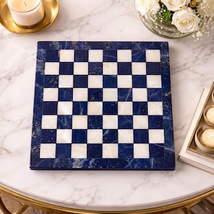 May include: A square chess board with a blue and white checkered pattern. The board is made of a dark blue stone with gold flecks and a white marble-like material for the squares. The board is on a white marble table.