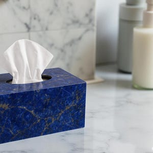 May include: A rectangular tissue box made of dark blue stone with gold veining. White tissues are pulled from the circular opening on top. The box is on a marble surface, with bottles in the background.