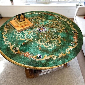May include: A round, green table with an ornate gold and cream-colored floral design. The table has a gold trim and sits on a wooden base. A book and decorative box rest on the table's surface.