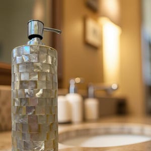May include: A decorative soap dispenser with a mosaic tile design. The dispenser is cylindrical with a silver pump and is made of iridescent, rectangular tiles in shades of white, beige, and gray. The dispenser is on a countertop.