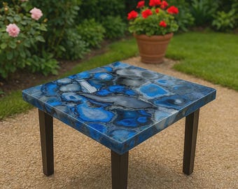 May include: Square side table with a blue and gray agate stone top. The table has a dark brown metal frame and legs. The table is outdoors on a gravel surface, with greenery and flowers in the background.