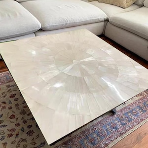 May include: Square coffee table with a light-colored, radial-patterned top. The table sits on a dark frame and is placed over a patterned rug and hardwood floor. A white sectional sofa is in the background.