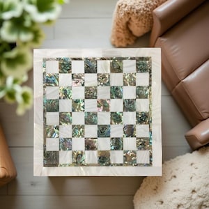 May include: Square chess board with a white border and a checkerboard pattern. The squares alternate between white and iridescent abalone shell. The board is set on a light-colored wooden surface.