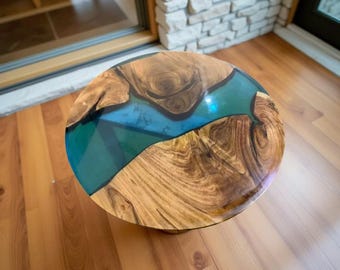 May include: Round wooden coffee table with a clear blue epoxy resin river design. The table features natural wood grain patterns in shades of brown and tan. A unique piece of furniture for a living room.