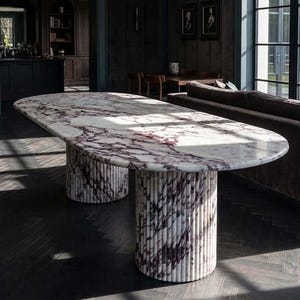 May include: An oval dining table featuring a white and deep purple veined marble top. The table is supported by two cylindrical, fluted marble pedestals that match the color and veining pattern of the tabletop surface, creating a cohesive look.