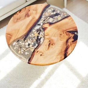 May include: Round wooden coffee table with a unique design. The table features a natural wood grain pattern with a central inlay of sand, pebbles, and starfish. The table is approximately 36 inches in diameter.