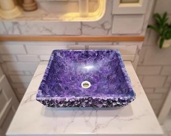 May include: A square, purple amethyst bathroom sink with a textured, crystalline edge. The sink has a central drain and sits on a white marble countertop. The sink is a unique, decorative element for a bathroom.