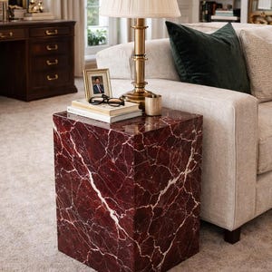 May include: A red marble side table with white veining. A brass lamp with a cream shade sits on top, along with books, a framed photo, and glasses. The table is next to a beige sofa.