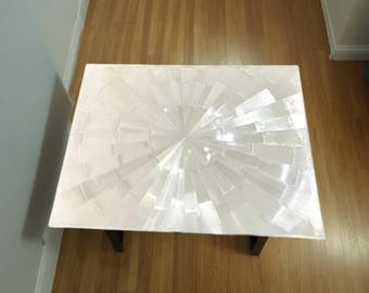 May include: Rectangular white coffee table with a radial design on the surface. The table has a dark base and is set on a hardwood floor. The tabletop is made of rectangular pieces arranged in a sunburst pattern.