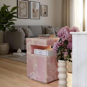 May include: A pink, rectangular side table with a unique, layered design. The table has a cutout section and is topped with a silver tray and a stack of books. A vase of purple flowers sits nearby, complementing the table's color. A gray sofa and framed art are in the background.