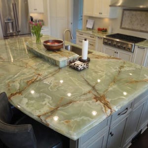 May include: A kitchen island with a green marble countertop featuring brown veining. The island has a built-in sink and a raised section. Cabinets are painted a light gray color. A wooden bowl with fruit sits on the countertop.