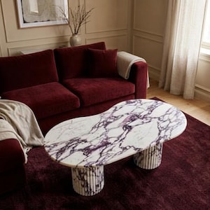 May include: A coffee table featuring an irregular, organic-shaped white marble top with prominent purple veining. The table is supported by two cylindrical, fluted marble bases that match the white and purple color pattern of the tabletop surface.