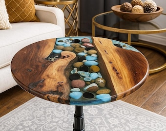 May include: A round wooden side table with a unique design. The tabletop features a river of blue resin with embedded stones, bordered by natural wood grain. The table stands 24 inches tall on a black, ornate pedestal base.