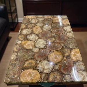 May include: A rectangular coffee table with a unique surface made of polished, multicolored stones. The stones are primarily shades of brown, tan, and gray, creating a natural, textured appearance. The table is set in a living room setting.