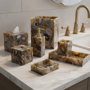 May include: A bathroom set featuring a tissue box, soap dispenser, wastebasket, soap dish, tray, and tumbler. The set is made of a brown and gray agate stone with gold accents. The items are arranged on a white marble countertop.