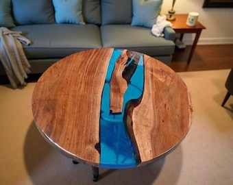 May include: Round wooden coffee table with a river of bright blue resin. The table has a natural wood grain finish and sits on black metal legs. The table's design incorporates a striking contrast between the wood and the resin.