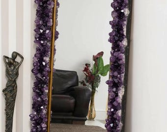 May include: A large rectangular mirror with a frame completely encrusted with deep purple amethyst crystals. The mirror has a gold-colored inner frame and a dark outer frame. The mirror is leaning against a wall.