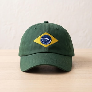 May include: A dark green baseball cap featuring an embroidered patch on the front. The patch displays a yellow diamond shape containing a blue circle with a white curved band and small white stars, representing the national flag of Brazil.
