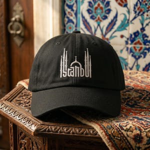 May include: A black baseball cap featuring white embroidery on the front panel. The design depicts a stylized architectural silhouette of a mosque with minarets and a central dome, with the text Istanbul integrated into the structure.
