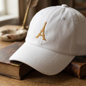 May include: White baseball cap featuring an embroidered gold Eiffel Tower design on the front. The cap has a curved brim and an adjustable strap at the back. A classic, casual accessory.