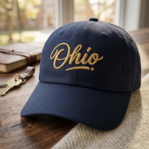 May include: A navy blue baseball cap featuring the word Ohio embroidered in gold script lettering across the front. The cap has a structured crown, a curved brim with tonal stitching, and a button at the top center of the fabric panels.