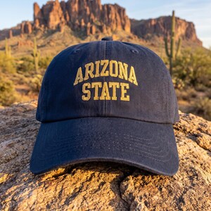 May include: A navy blue baseball cap featuring the text ARIZONA STATE embroidered in gold thread across the front. The cap has a structured crown, a curved brim with visible stitching, and a button at the top center of the fabric dome.