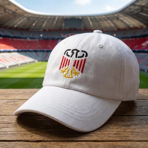 May include: A white baseball cap featuring an embroidered stylized eagle emblem on the front panel. The embroidery uses black, red, and yellow thread to depict the eagle design. The cap has a curved brim and a structured crown with ventilation eyelets.