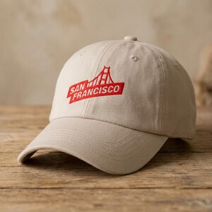 May include: A beige baseball cap with a curved brim. The cap features an embroidered red design of the Golden Gate Bridge and the words "San Francisco" in a red rectangular box. The cap is made of a soft fabric.