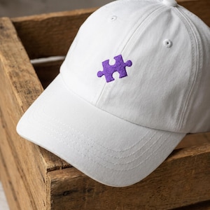 May include: White baseball cap with a purple puzzle piece embroidered on the front. The cap is made of a soft fabric and has a curved brim. The cap is sitting inside a wooden box.