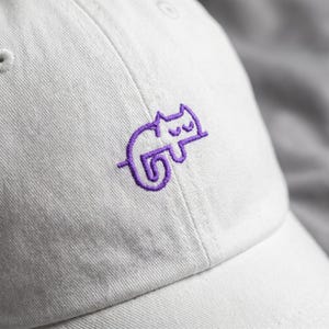 May include: A white baseball cap with a purple embroidered cat design on the front. The cat is stylized and appears to be sleeping. The cap is made of a soft fabric and has a curved brim.