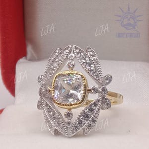 May include: A gold and silver ring with a large, square, clear gemstone at the center. The gemstone is surrounded by a gold setting and a silver filigree design. The ring is set against a white background.
