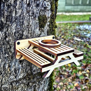 Può includere: Un mangiatoia per uccelli in legno a forma di tavolo da picnic, montata su un tronco d'albero. La mangiatoia ha una piccola ciotola per il cibo e due panchine.